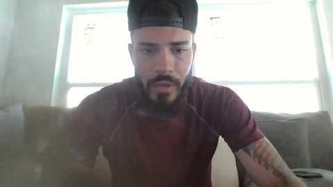 Media: Video of a bearded man in a black backward cap, maroon shirt, and headphones, sitting on a gray couch, looking at a phone.