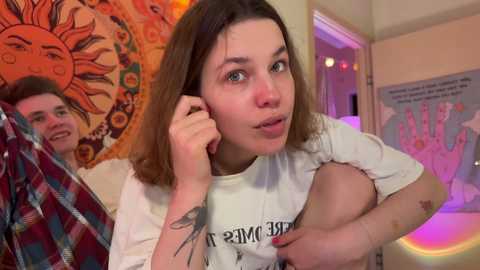 Media: Video of a young woman with shoulder-length brown hair, wearing a white shirt, holding a phone, in a room with a sunflower tapestry, plaid blanket, and posters.