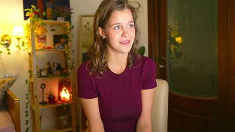 Media: Video of a young Caucasian woman with shoulder-length brown hair, wearing a purple T-shirt, smiling thoughtfully in a warmly lit, cozy living room with bookshelves, candles, and floral decorations.