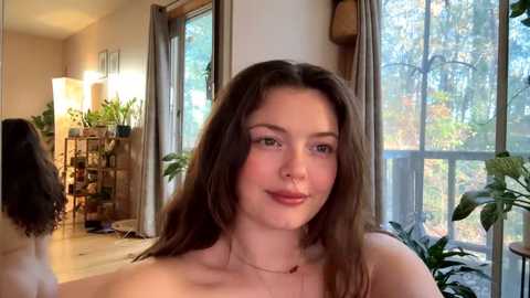 Media: Video of a young, topless white woman with long brown hair, wearing a nose ring, smiling in a cozy room with large windows, potted plants, and wooden furniture.