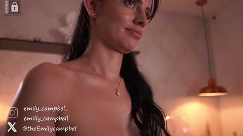 Media: Video of a topless woman with long black hair, fair skin, and a confident expression, wearing a necklace. Warm, ambient lighting in a modern, dimly lit room with a red pendant light and a shelf.