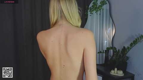 Media: Video of a nude blonde woman with fair skin, viewed from behind, standing in a dimly lit room with a mirror, potted plants, and candles on a table.