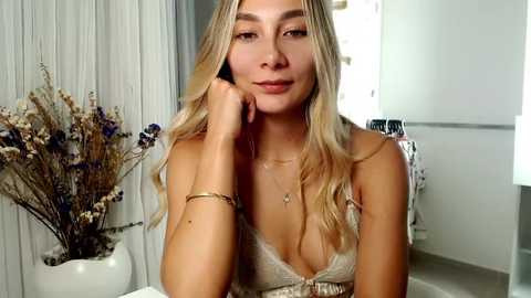 Media: Video of a young woman with long blonde hair, fair skin, and light makeup, wearing a gold lace top, resting her chin on her hand. Background features white curtains, a vase of dried flowers, and a partially visible mirror.