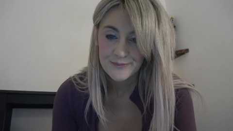 Media: A video of a blonde woman with straight hair, light skin, and blue eyes, wearing a purple top, smiling slightly. She's indoors against a plain, light-colored wall.