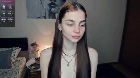 Media: A video of a young, pale-skinned woman with long, straight brown hair, wearing a necklace and a sleeveless top, standing in a dimly lit bedroom with floral-patterned bedding and a framed black-and-white photo on the wall.