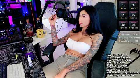 Media: Video of a tattooed woman with long dark hair, wearing a white strapless top and beige shorts, sitting in a gaming chair. Background features a messy desk with computer peripherals, bottles, and a keyboard. Live-streaming screen overlays show viewers and chat.