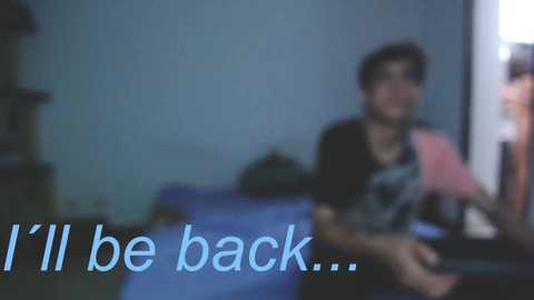 Media: Video of a young person with short dark hair wearing glasses and a black T-shirt, sitting on a bed in a dimly lit room. Text on the image reads, \"I'll be back...\" in light blue font.