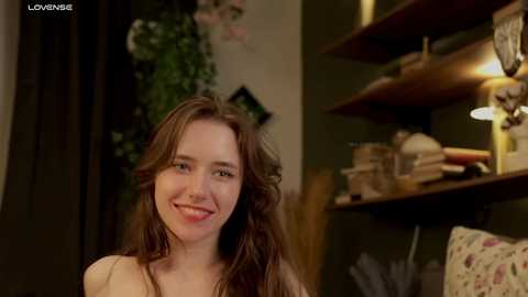 Media: Video of a smiling young woman with long brown hair and fair skin, wearing a light-colored top, in a cozy, dimly-lit room with wooden shelves and a lamp.