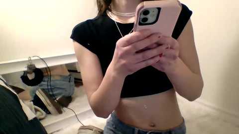 Media: A young woman with fair skin, small breasts, and a slender build takes a selfie in a cluttered bedroom, wearing a black crop top and blue jeans.