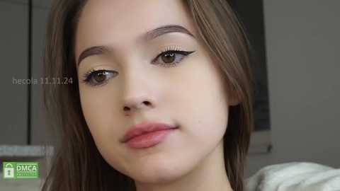 Media: Video of a young woman with fair skin, straight brown hair, and light makeup, including mascara and light pink lipstick, in a neutral indoor setting.