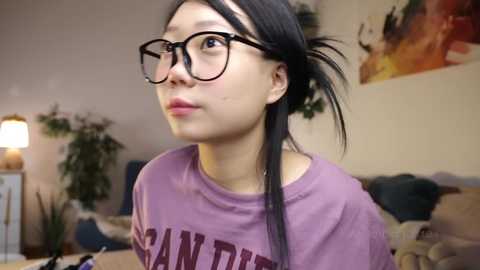 Media: Video of an Asian woman with long black hair, wearing black-framed glasses, a purple shirt, and a neutral expression. The background features a beige wall, a potted plant, and a colorful abstract painting.