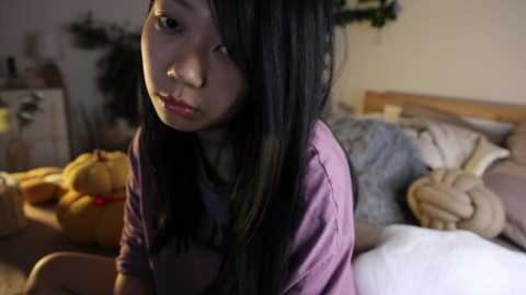 Media: Video of a young Asian girl with long black hair, wearing a purple shirt, sitting on a bed in a dimly lit room with stuffed animals and decorative pillows.