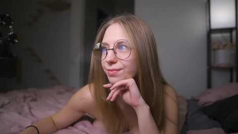 Media: Video of a slender, light-skinned young woman with long blonde hair and round glasses, resting on her elbows on a bed with pink sheets, in a dimly lit bedroom.