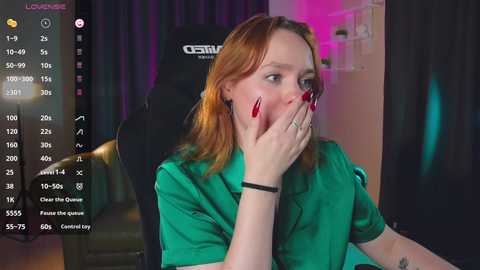 Media: Video of a young woman with fair skin, red hair, and green shirt, covering her mouth with her hands in surprise, sitting in a black gaming chair.