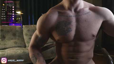 Media: Video of a shirtless, muscular man with tattoos on his chest and arms, standing in a dimly-lit living room with a floral-patterned couch and dark curtains.