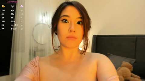 Media: A video of a young Asian woman with light skin and dark brown hair in a casual, intimate bedroom setting. She has a surprised expression, wearing a light pink top.