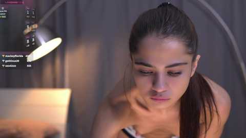 Media: Video of a topless young woman with fair skin and dark hair in a ponytail, focused on a laptop in a dimly lit room with a hanging lamp and grey curtains.