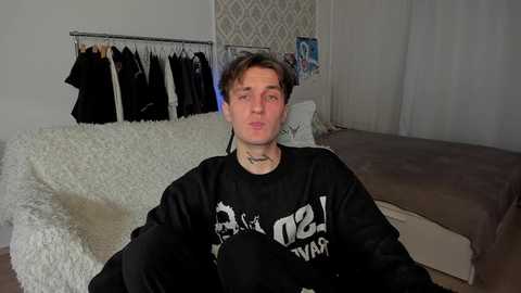 Media: Video of a young man with short, dark hair, wearing a black shirt with white text, sitting on a beige bed with a white fur blanket. Background includes a metal clothing rack with black clothes, white wallpaper, and sheer curtains.