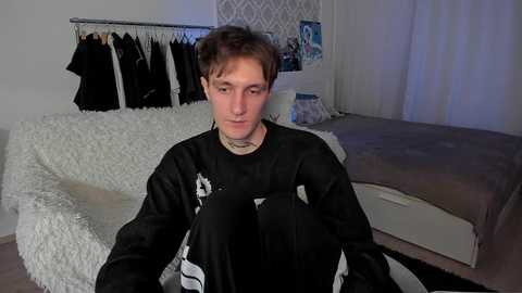 Media: Video of a young man with short brown hair, wearing a black sweatshirt, sitting on a bed in a minimalist bedroom with white curtains and a hanging rack of clothes.