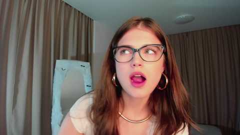 Media: Video of a Caucasian woman with long brown hair, wearing glasses, hoop earrings, and a white top, standing in a beige room with curtains. She has an open-mouthed expression.