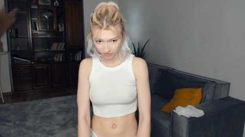 Media: Video of a blonde woman with a slim physique wearing a white crop top, standing in a modern living room with a grey sofa and bookshelf in the background.