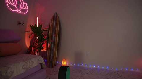 Media: Video of a dimly-lit bedroom with a bed, a potted plant, a lit candle, a neon lotus, and a surfboard leaning against the wall.