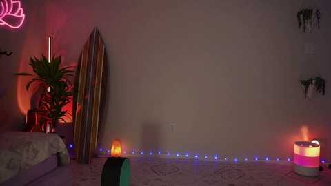 Media: Video of a dimly lit room with a beige wall, surfboard leaning against it, potted plants, neon lights, and glowing candles on the floor.