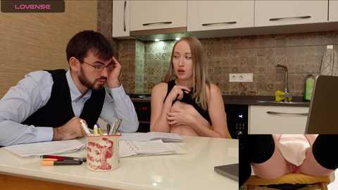 Media: Video of a man in a vest and glasses, and a blonde woman in a black top, sitting at a kitchen table. Close-up inset shows her exposed buttocks.