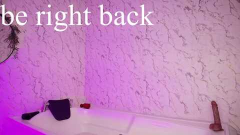 Media: Video of a white marble bathroom with a bathtub, purple lighting, a black towel, a red candle, and a wooden dildo. Text reads, \"be right back.\