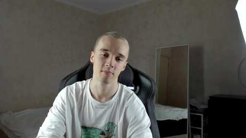 Media: A video of a young Caucasian man with a shaved head, wearing a white graphic T-shirt, sitting in a black gaming chair, in a dimly lit, plain bedroom with a mirror and a bed visible in the background.