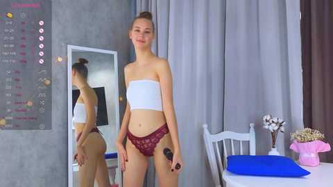 Media: Video of a slim, light-skinned young woman with auburn hair in a bun, wearing a white bandeau top and maroon lace panties, standing in a modern, minimalistic bedroom with gray walls, a mirror, and a white chair.