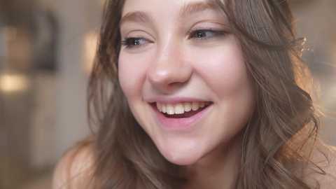 Media: Video of a young Caucasian woman with light skin, long wavy brown hair, and blue eyes, smiling broadly, her teeth visible. The blurred background suggests an indoor setting.