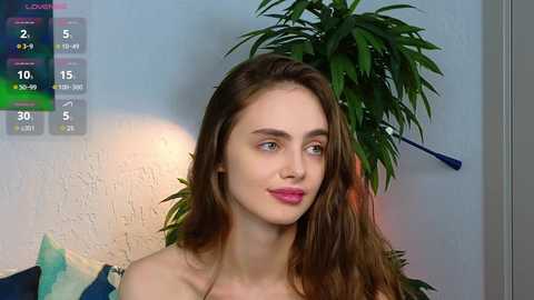 Media: A video of a young, fair-skinned woman with long, wavy brown hair, wearing pink lipstick, sitting indoors next to a green potted plant. The background features a white wall and a digital clock displaying 5:30.