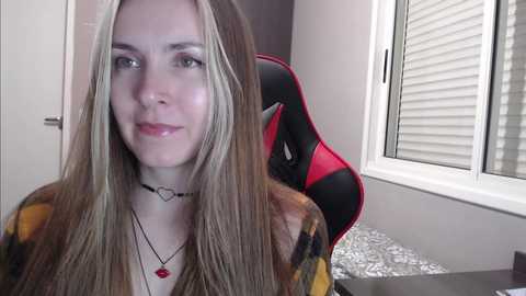 Media: A video of a young, fair-skinned woman with long, blonde hair, wearing a red necklace and a plaid jacket, sitting in a gaming chair in a modern, brightly-lit room.