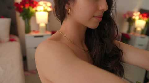 Media: Video of a light-skinned woman with long, dark hair, wearing a gold necklace, smiling, adjusting her hair. Background shows a white dresser with red flowers and a lamp.