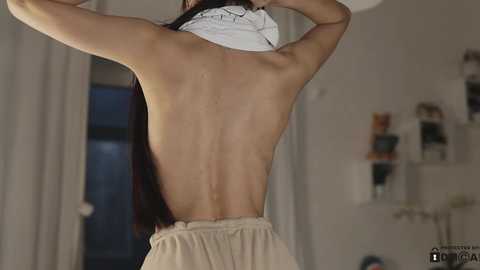 Media: Video of a topless woman with long dark hair, wearing beige shorts, lifting a white shirt. Background features a modern, minimalistic room with shelves and plants.