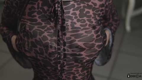 Media: Video of a person with medium skin tone wearing a tight, sheer leopard print dress with a black lace trim, revealing their breasts and nipples. The background is blurred, showing a tiled floor.