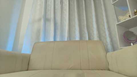 Media: Video of a light-colored, cushioned couch with a soft, cream fabric, facing a white curtain. To the right, a white bookshelf with decorative items is visible. The setting appears clean and minimalist.