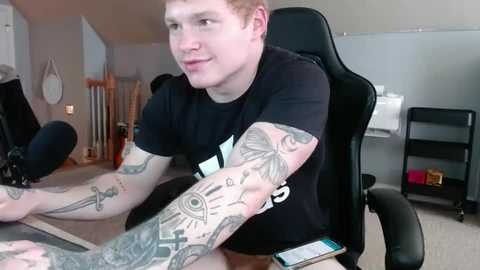 Media: Video of a young man with fair skin, short red hair, wearing a black t-shirt, seated in a black gaming chair, with numerous tattoos on his arms, in a modern, cluttered room with a desk, microphone, and a wall-mounted whiteboard.