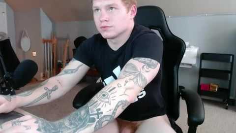 Media: Video of a young, fair-skinned man with red hair, wearing a black T-shirt, seated in a black gaming chair, showcasing extensive arm tattoos in a dimly lit, cluttered room with a computer setup.