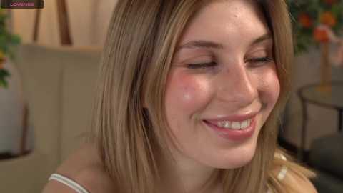 Media: Video of a young Caucasian woman with fair skin and blonde hair, smiling with closed eyes, wearing a white top, in a warmly lit room with potted plants in the background.