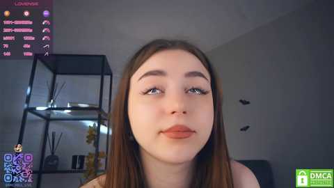 Media: Video of a young woman with long brown hair, fair skin, and light makeup, captured in a modern, minimalistic room with a glass shelving unit and decorative elements.