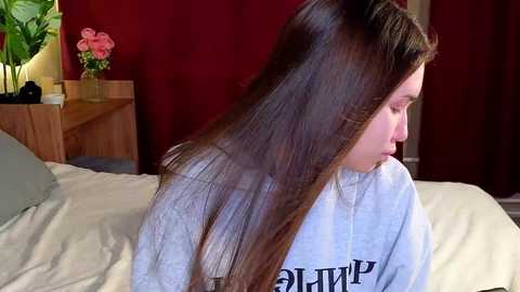 Media: Video of a young woman with long, straight brown hair, sitting on a bed with a beige duvet. She wears a light grey hoodie. The room features red curtains and a wooden nightstand with a vase of pink flowers.