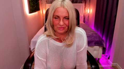 Media: Video of a blonde, middle-aged woman with a medium build, wearing a white, sheer top, sitting on a chair in a dimly lit, cozy room with purple lighting and a bed in the background.