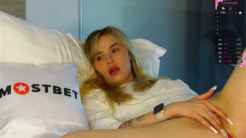 Media: Video of a blonde woman with fair skin, wearing a white sweater, lying on a bed with white pillows and a \"MOSTBET\" pillow, looking at a phone.