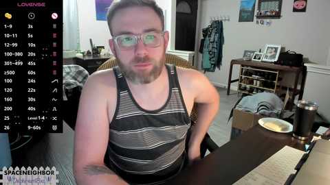 Media: Video of a bearded, pale-skinned man in glasses, wearing a striped tank top, seated at a messy desk in a cluttered bedroom.