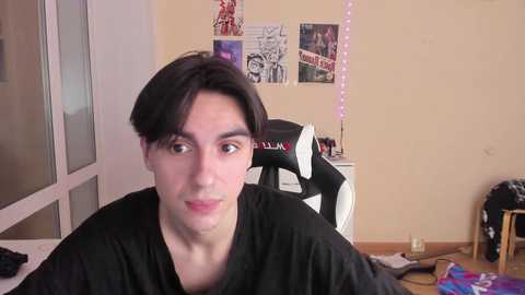 Media: Video of a young man with dark hair, wearing a black shirt, seated in a gaming chair in a cluttered bedroom with posters, fairy lights, and a desk.