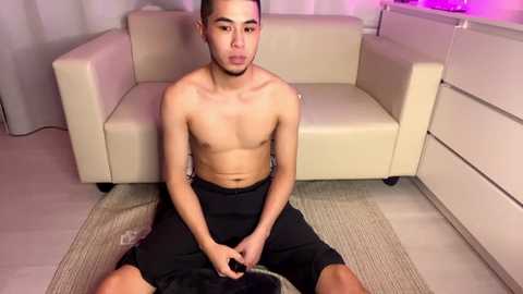 Media: Video of a shirtless Asian man with short black hair, sitting on a beige carpet in a modern living room with white furniture and purple lighting.