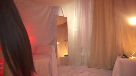 Media: Video of a dimly lit, minimalist room with sheer white curtains, a mirror, and a lamp casting soft light.