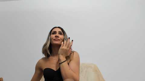 Media: Video of a slender, light-skinned woman with shoulder-length, dark brown hair, wearing a black strapless top, smiling and holding her hand to her mouth, against a plain white background.